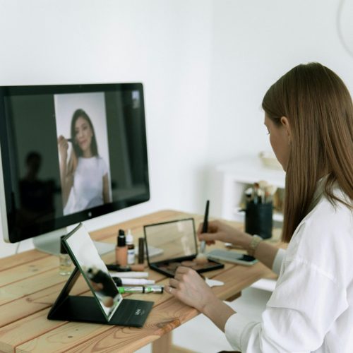 A woman follows an online makeup tutorial using a computer and tablet at home.